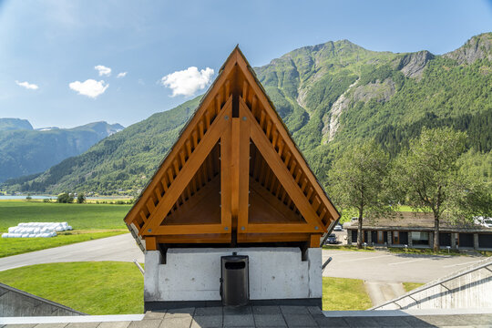 View of a wooden triangular structure with a trash can below it, set against a backdrop of green fields, distant mountains, and a bright sky, Fjaerland, Norway.