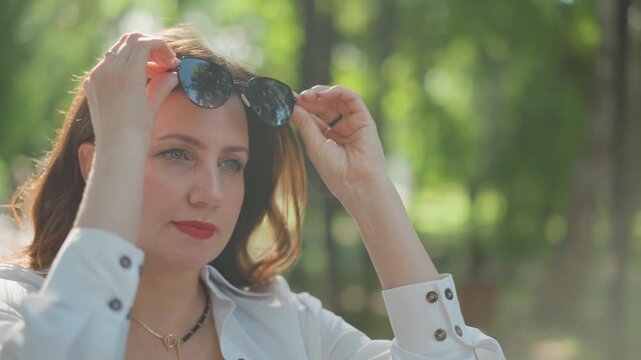Caucasian woman adjusting sunglasses park, closeup fashion portrait, red lipstick and confident gaze, backlit bokeh and urban shade, assertive mood, chic accessory focus, lifestyle decree tone