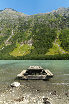 View of a picnic table submerged in the turquoise waters of a glacial lake, framed by rugged mountains and lush green patches, Boyabreen glacier, Fjaerland, Norway.