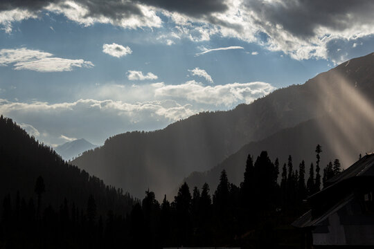 View of sun rays piercing through dramatic clouds over the dark, silhouetted mountains and trees in a stunning natural display, Arang Kel, Pakistan.