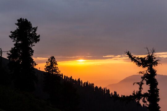 View of the sun sinking behind silhouetted trees on a mountain ridge with a warm glow illuminating the sky, Pir Chanasi, Azad Kashmir, Pakistan.