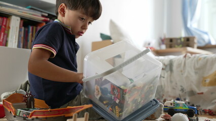 Young boy lifting plastic container filled with colorful toys, preparing for imaginative play,...