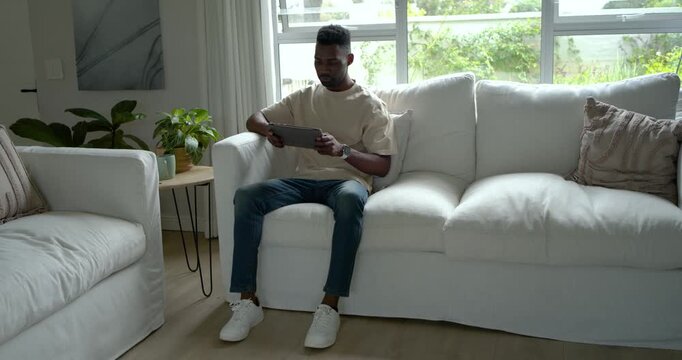 African American man sitting on white sofa in living room holding tablet and watching funny clip