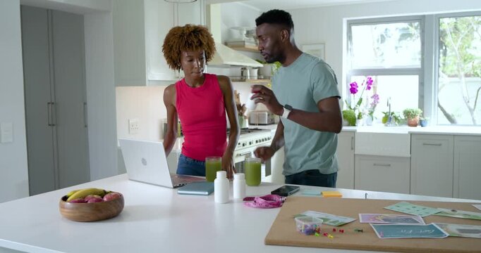 Diverse couple sipping green drinks, picking up white bottle and arranging craft board using laptop
