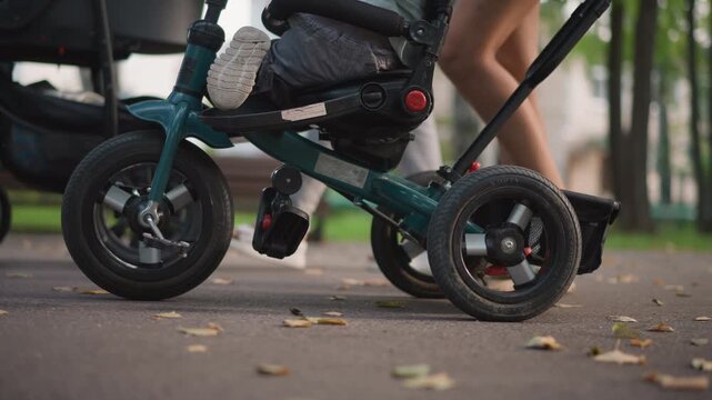 Closeup of stroller and tricycle wheels rolling on park pavement, focus on tire tread, pedal and sneaker movement, subtle autumn leaves and textured asphalt, everyday mobility detail conveying active
