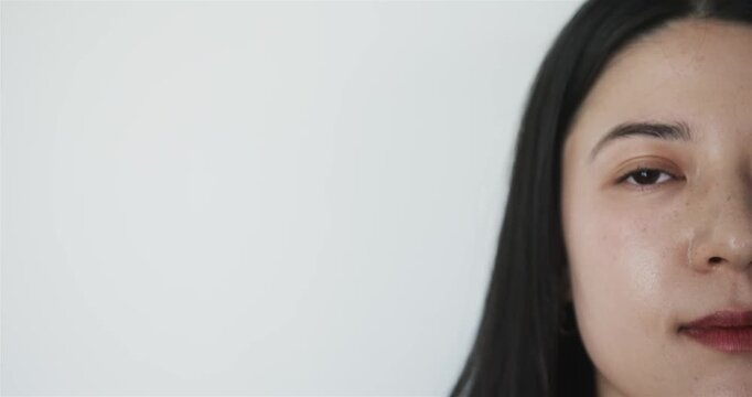 Asian woman slowly opening eye from closed state and smiling on plain pale backdrop, copy space