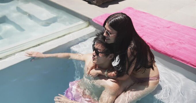 Diverse couple in swimwear straddling, holding while man pointing after spotting near pool steps