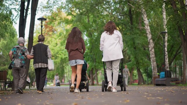 Two white women walking with stroller through busy community park, multigenerational neighbors passing by, benches and tree canopy framing pathway, inclusive neighborhood lifestyle scene with ambient