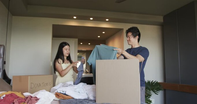 Asian couple woman picking through pile while man holding teal shirt at home, filling donations box