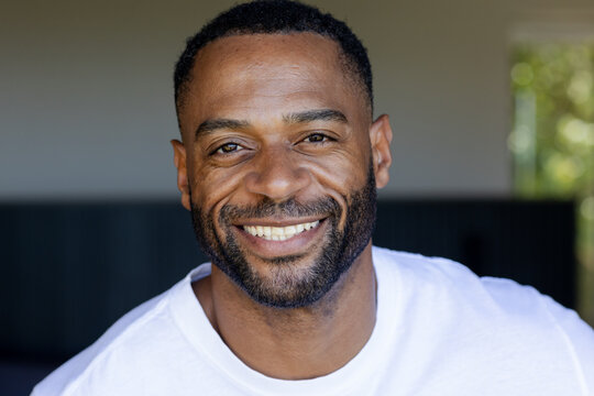 African American man smiling while wearing plain white T-shirt on shaded porch, sunlight filtering