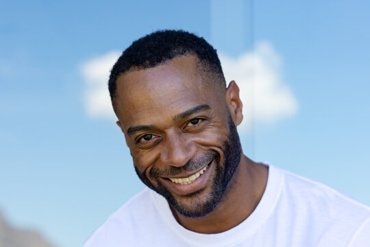 Mature African American man smiling at camera outdoors by reflective glass panel wearing white tee