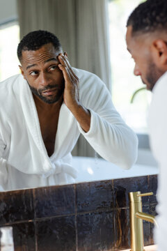 Mature African American man wearing white robe, patting wet face at vanity mirror near gold faucet