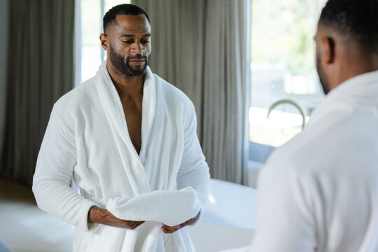 Mature African American man standing in bathroom wearing bathrobe holding white towel facing mirror