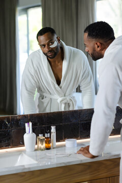 Middle-aged African American man in bathrobe leaning at bathroom mirror inspecting under-eye patch
