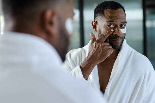 Mature African American man in white bathrobe at mirror, applying crescent under-eye patches