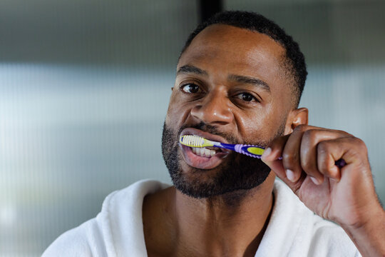 African man brushing teeth with blue-yellow toothbrush in white bathrobe in bath near ribbed glass