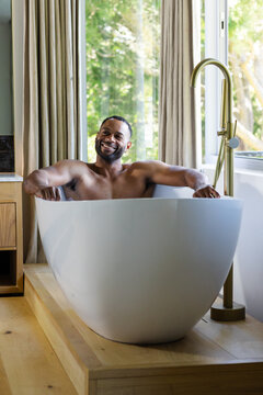 Mature African American man barechested sitting in oval tub on deck smiling, brass-faucet, curtains