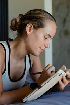 Female reclining on bed, writing in bound notebook, wearing smartwatch and gold hoop earrings