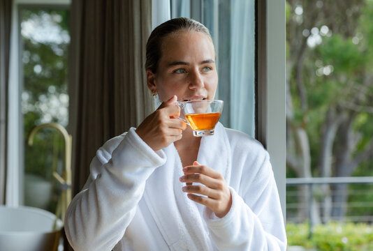 Woman standing near glass door sipping tea from clear cup in white bathrobe beside bathtub