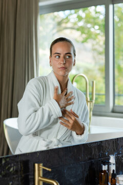 Woman standing at bathroom vanity adjusting white bathrobe beside window with freestanding bathtub
