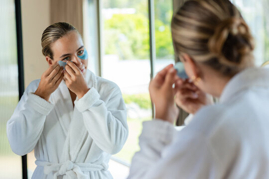 Female standing at mirror in home bathroom applying blue under-eye gel patches wearing white robe