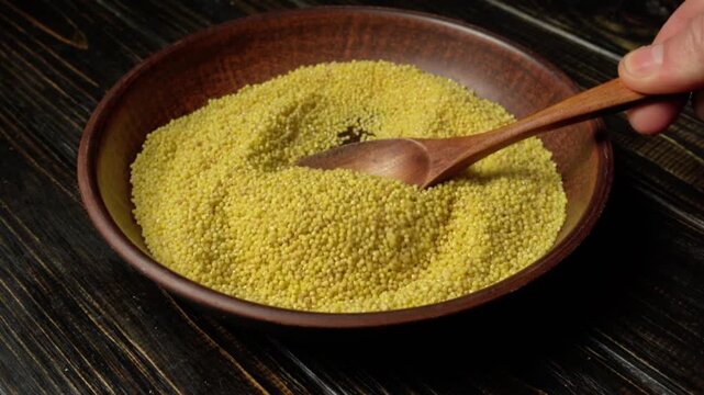 Hand scooping yellow millet grain from a wooden bowl with a wooden spoon against a dark wooden background in a kitchen setting