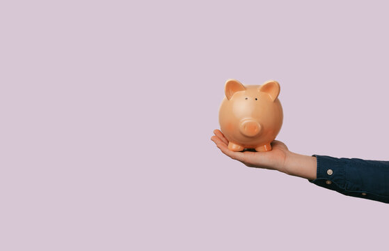 Hand Holding Ceramic Piggy Bank On Pastel Pink Background. Minimal Studio Composition Evoking Financial