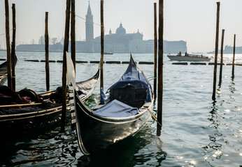 Obraz premium Venice, Italy - February 04, 2024: Gondola moored beside wooden poles in a tranquil lagoon with historic buildings and a bell tower visible in the background