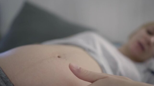 Close up of a resting pregnant woman gently stroking her belly while lying down on the sofa at home