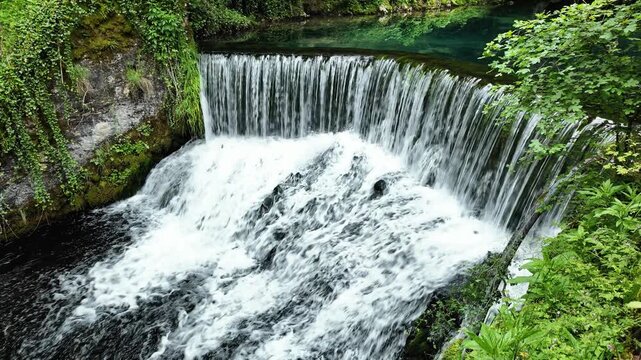 Water flowing over a wide natural rock ledge forming a beautiful waterfall surrounded by lush green forest and clear turquoise river. Pure nature, freshwater resources, travel destination relaxation