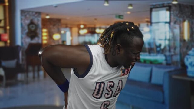 Man holds lower back and winces while wearing usa basketball jersey with flag patch in a hotel lobby near sofa and lamp; pain backache.