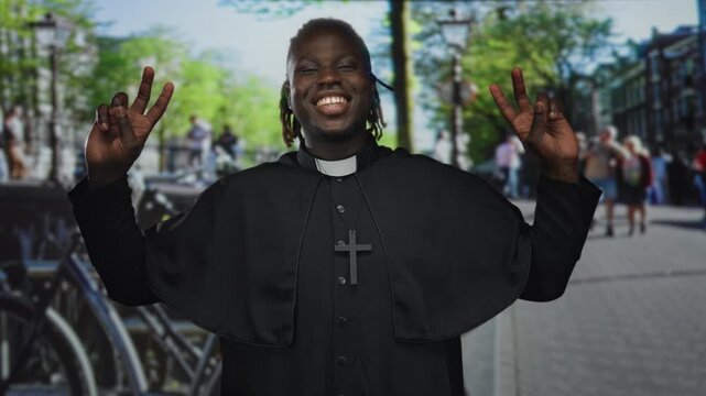Man priest in cassock holds up peace sign with both hands on a busy street; joy faith community hope.