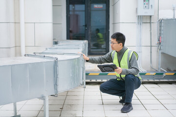 Engineer crouching to inspect ventilation ducts while holding a tablet computer