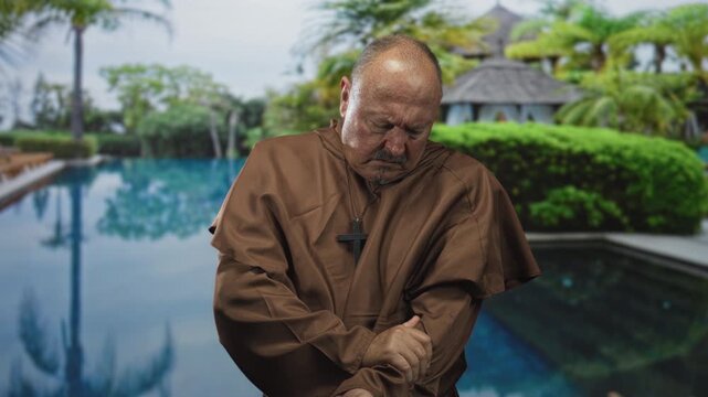 Man clutching arm in brown robe and cross necklace beside resort pool near a thatched building; solitude contemplation.