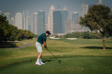Fototapeta premium Male golfer in green shirt and white shorts prepares to swing golf club on tee box with city skyline and green fairway in the background