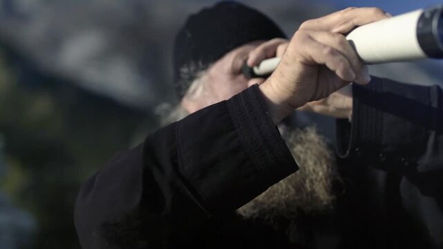 Elderly orthodox monk with a long beard observing the landscape with a spyglass in Mount Athos