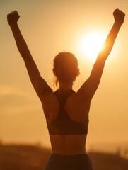 Fototapeta premium A woman in a sports bra and workout attire celebrating with fists raised against a sunset background.