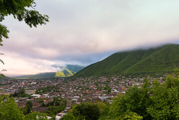 Obraz premium Shaki City, Azerbaijan, nestled in a valley with green mountains partially covered by atmospheric clouds. Overhead view of houses and trees under a soft sky