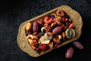 Dried tropical fruits, nuts, and raisins in a golden bowl on black stone table.