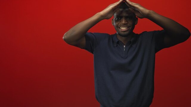 Young black man smiling, wearing glasses and navy polo, hands forming a triangle above his head in a red studio; joy unity.