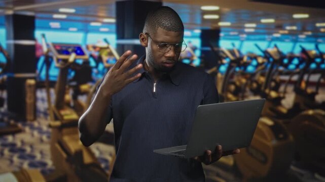 Man holding laptop with left hand, right fingers splayed near head while reading screen in a gym building; concentration remote coaching.