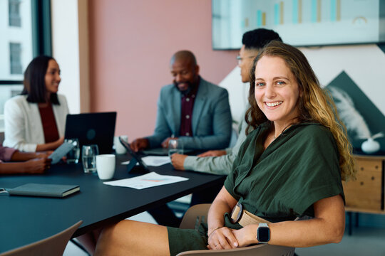 Young businesswoman in smart casual smiling at camera while colleagues discuss project in boardroom