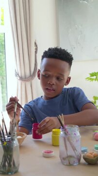Vertical video: Arranging brushes, boy wearing blue tee painting eggs at home table with glass jar