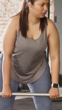 Vertical video: Gripping barbell, woman in gray tank performing bent-over rows at gym for strength