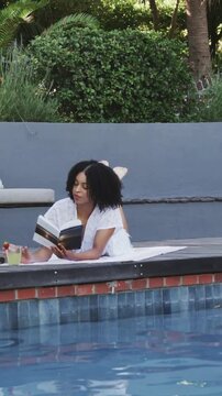 Vertical video: Reading woman in white coverup on towel poolside, grabbing book, drink, sun warming