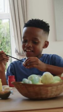 Preteen boy painting egg at table by window light holding brush, paints, bowl, smiling frame 12