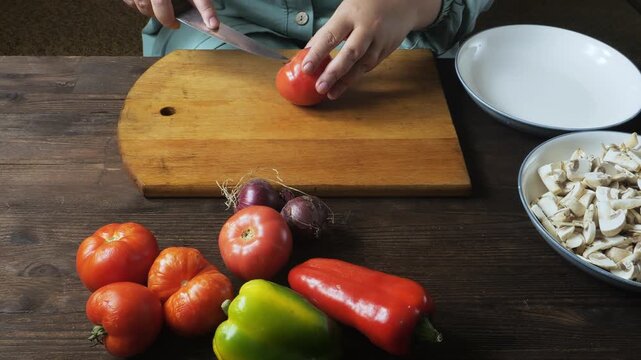A woman's hands cut fresh red tomatoes with a knife on a cutting board on a wooden table. Medium shot.