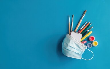 Colorful pencils and paints in a medical face mask on a blue background viewed from above