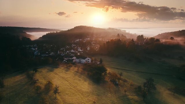 Flying over a rural landscape with hills, meadows and woods, with a village and illuminated fog at sunrise. Aerial footage with sideways motion.