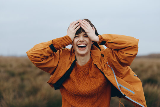 Happy woman in orange jacket and sweater laughs outdoors in nature, holding her head with both hands. Concept of woman lifestyle, joy, freedom, and autumn fashion.